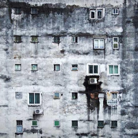 A weathered multi-story building facade with numerous windows, some featuring air conditioners, showing signs of aging and discoloration.