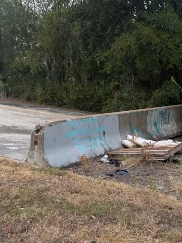 A concrete roadside barrier with graffiti, white bags, wooden planks, and debris on dry grass near a road with trees in the background.