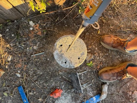 Power drill drilling into concrete disc with three holes, brown work boots, and tools on dirt ground near wooden fence.