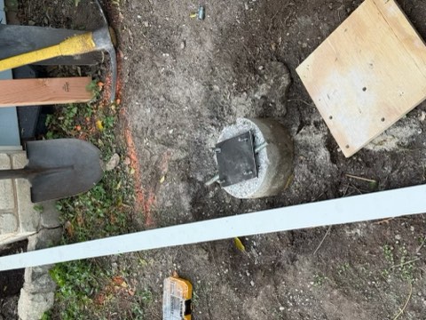 Concrete post with metal plate, wooden board, shovel, hammer, and tools on dirt ground.
