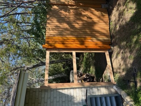 Wooden deck with slatted roof over stone wall, surrounded by trees and stacked firewood.
