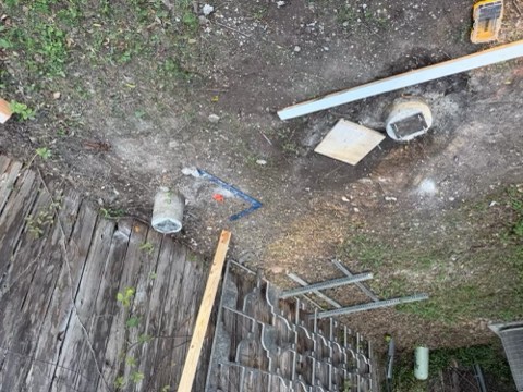 Overhead view of ground with wooden deck, concrete pipes, blue L-tool, white plank, square wood, round concrete object, metal brackets, dirt, grass, and yellow tool.
