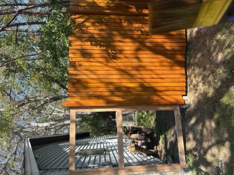 Wooden slatted structure elevated on posts, tree branches with green leaves above, dirt ground, and metal roof section below.