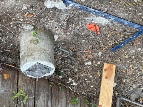 Concrete cylinder with metal plate and small plants, blue metal frame, orange tag marked '5', wooden plank, and wooden deck on dirt ground with rocks and debris.