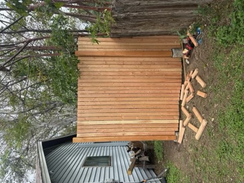 Wooden shed with slatted walls next to a gray metal building with a window; stacked wood planks and tools on ground near fence and trees.