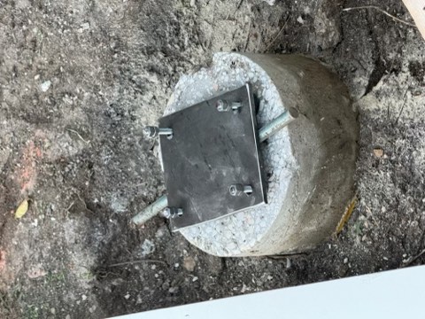 Concrete cylinder with metal plate secured by bolts, set in dirt ground.