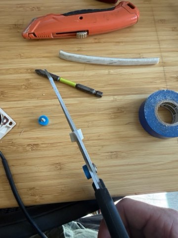 Hand holding scissors over wooden surface with orange utility knife, blue tape, screwdriver, and small blue component.