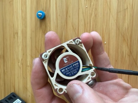 Hand holding a small computer fan with labeled sticker, wires connected, blue screw on wooden surface.