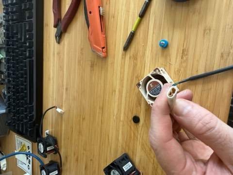 Hand holding a small fan with wires, on a wooden surface with tools and keyboard nearby.