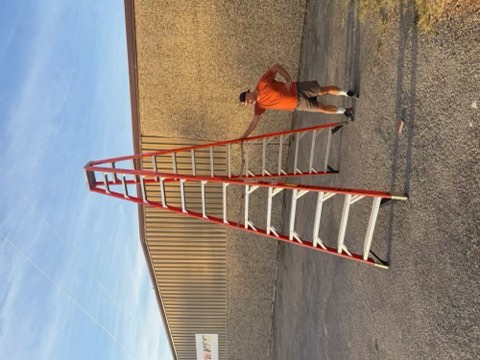 Man in orange shirt stands next to red and white ladder on gravel near building with blue sky.
