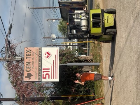 A man stands near a sign for Cen-Tex 511, with a yellow forklift on a gravel road and utility poles in the background.