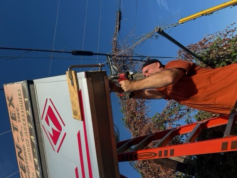 Man in orange shirt uses power drill on sign attached to ladder under blue sky.