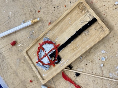 A wooden tray with a black stripe and red and gray materials sits on a wooden surface, surrounded by paintbrushes and paint splatters.