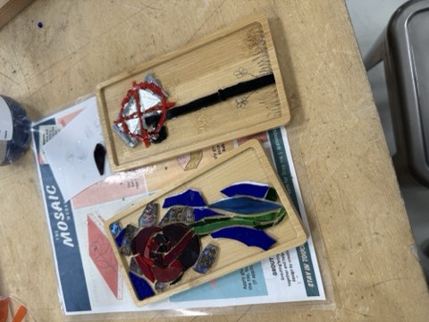 Two mosaic artworks with colored glass pieces on wooden trays rest on a magazine titled "MOSAIC" on a wooden surface, with a small container nearby.