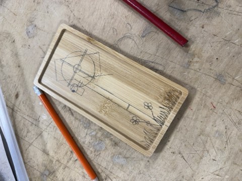 A wooden board with hand-drawn geometric shapes and floral designs, accompanied by a red pencil and an orange pencil, placed on a scratched wooden surface.
