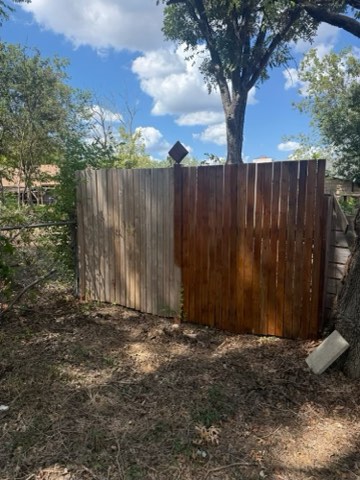 A wooden fence with light and dark brown planks, a tree with a diamond-shaped sign, and a blue sky with clouds in the background, with dry ground and other trees nearby.