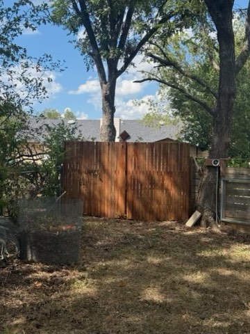 A wooden fence stands in a yard with trees, a stone structure, and a house roof visible in the background under a partly cloudy sky.
