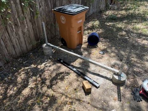 Outdoor yard with wooden fence, orange trash bin, blue bucket, metal rails, concrete bases, and tools on dirt ground.