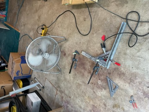 Workshop floor with fan, cardboard boxes, measuring tools, and cables scattered around.