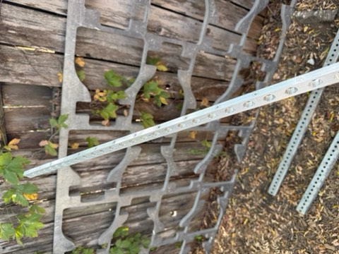 Metal grid structure with perforated bars leaning against weathered wooden fence, surrounded by fallen leaves and green foliage.