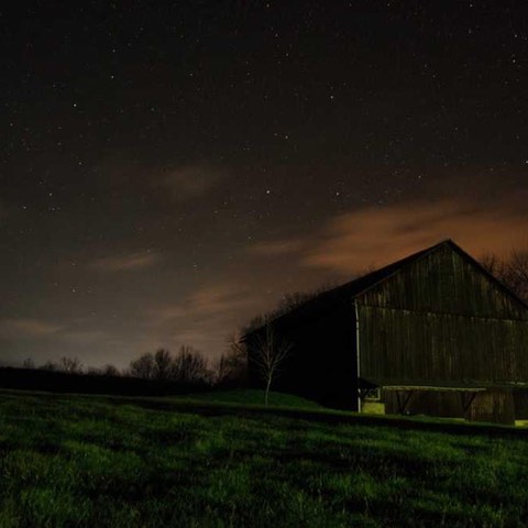 A dark barn stands on a grassy field under a starry night sky with faint clouds.
